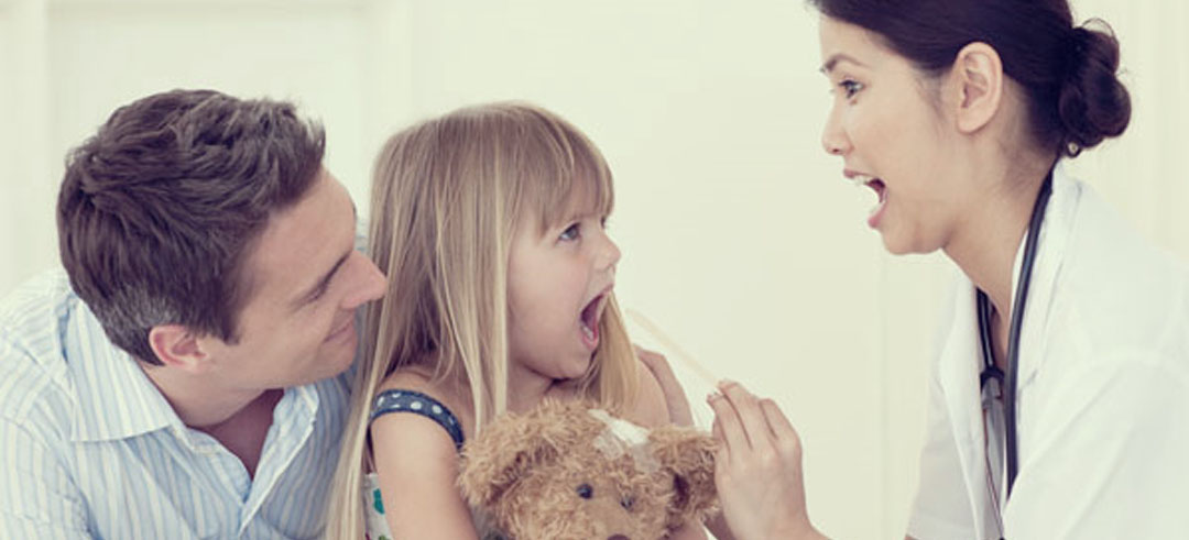 Mujer médico revisando la boca de una niña con una paleta mientras el padre está detrás